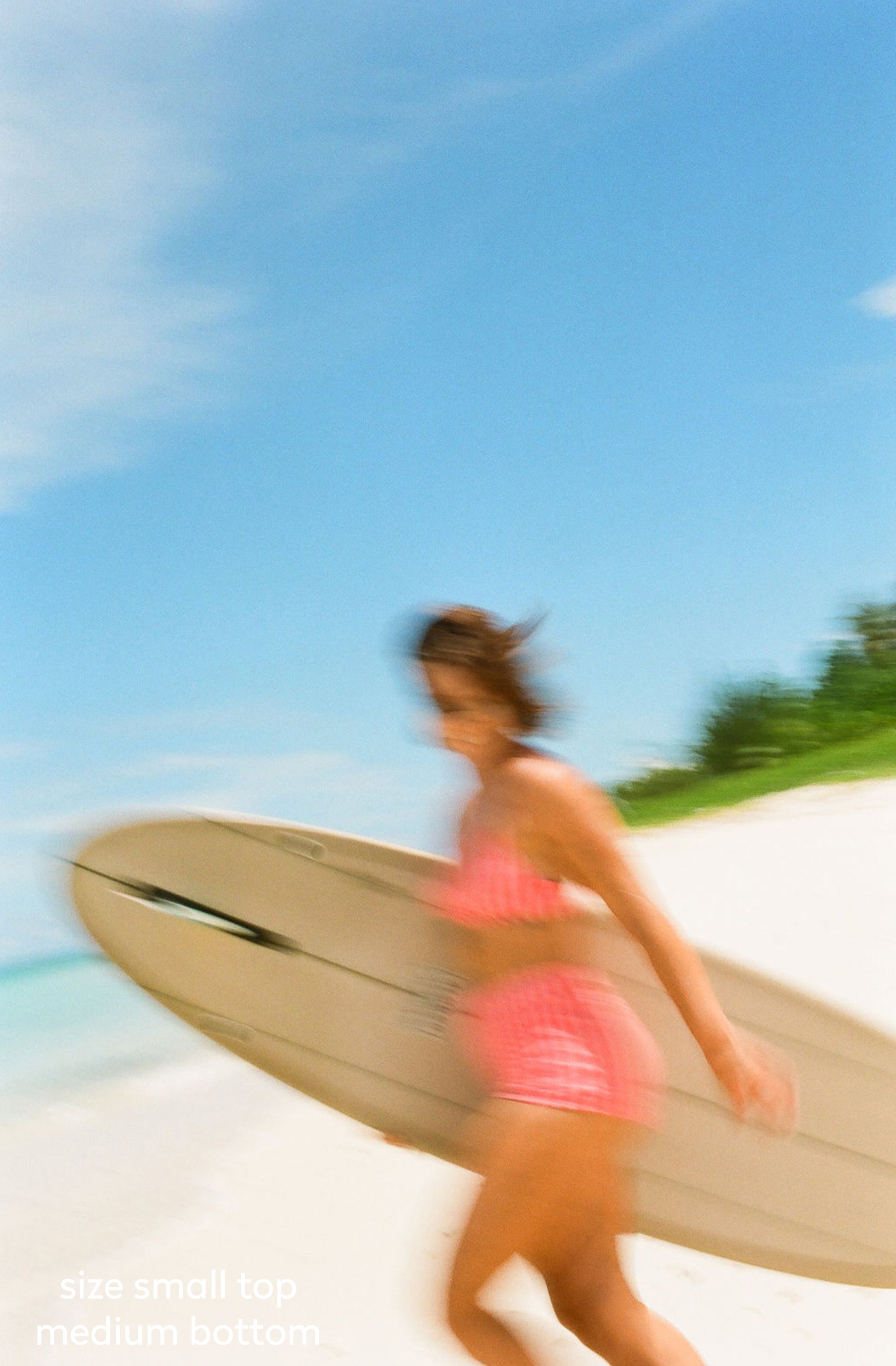 A woman carrying a large surfboard on a sunny beach, wearing a pink bikini. The image has a strong motion blur effect, and the background shows a bright blue sky and white sand.
