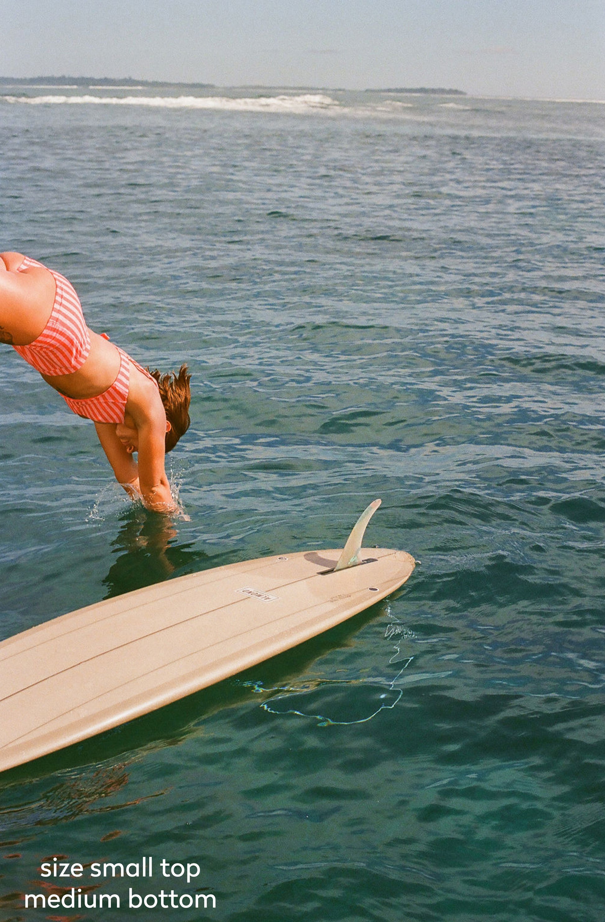 A person in a pink striped bikini dives headfirst into the ocean next to a light-colored surfboard. The image has a text overlay at the bottom left reading 'size small top medium bottom'.