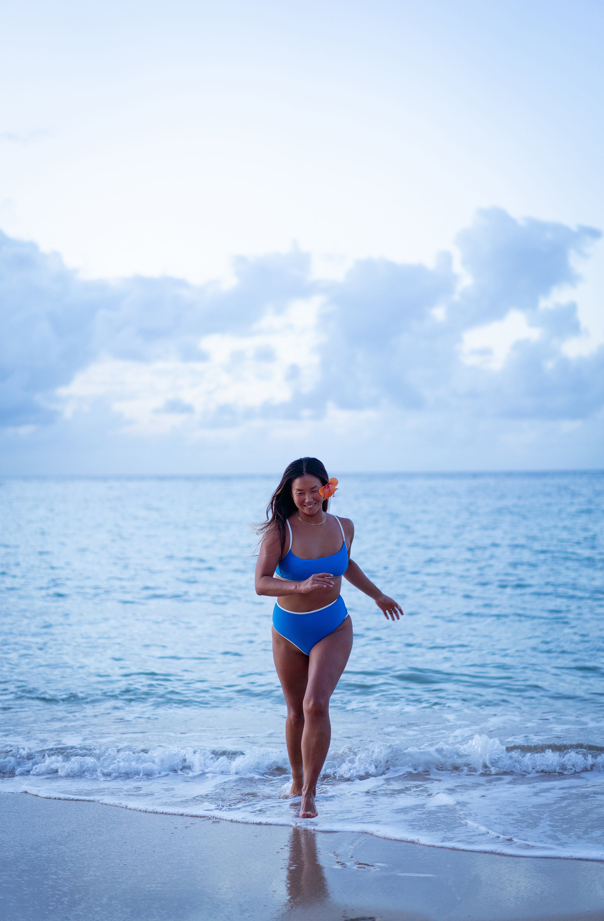 A woman in a blue two-piece swimsuit walks out of the ocean onto a sandy beach under a cloudy sky.