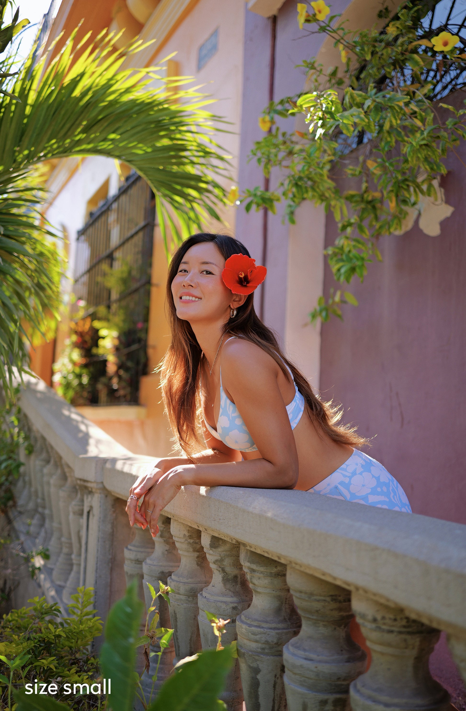 A woman in a white bikini top and light blue patterned skirt leans on a stone balcony railing, with a red flower in her hair, set against a warm-toned building wall and green foliage.