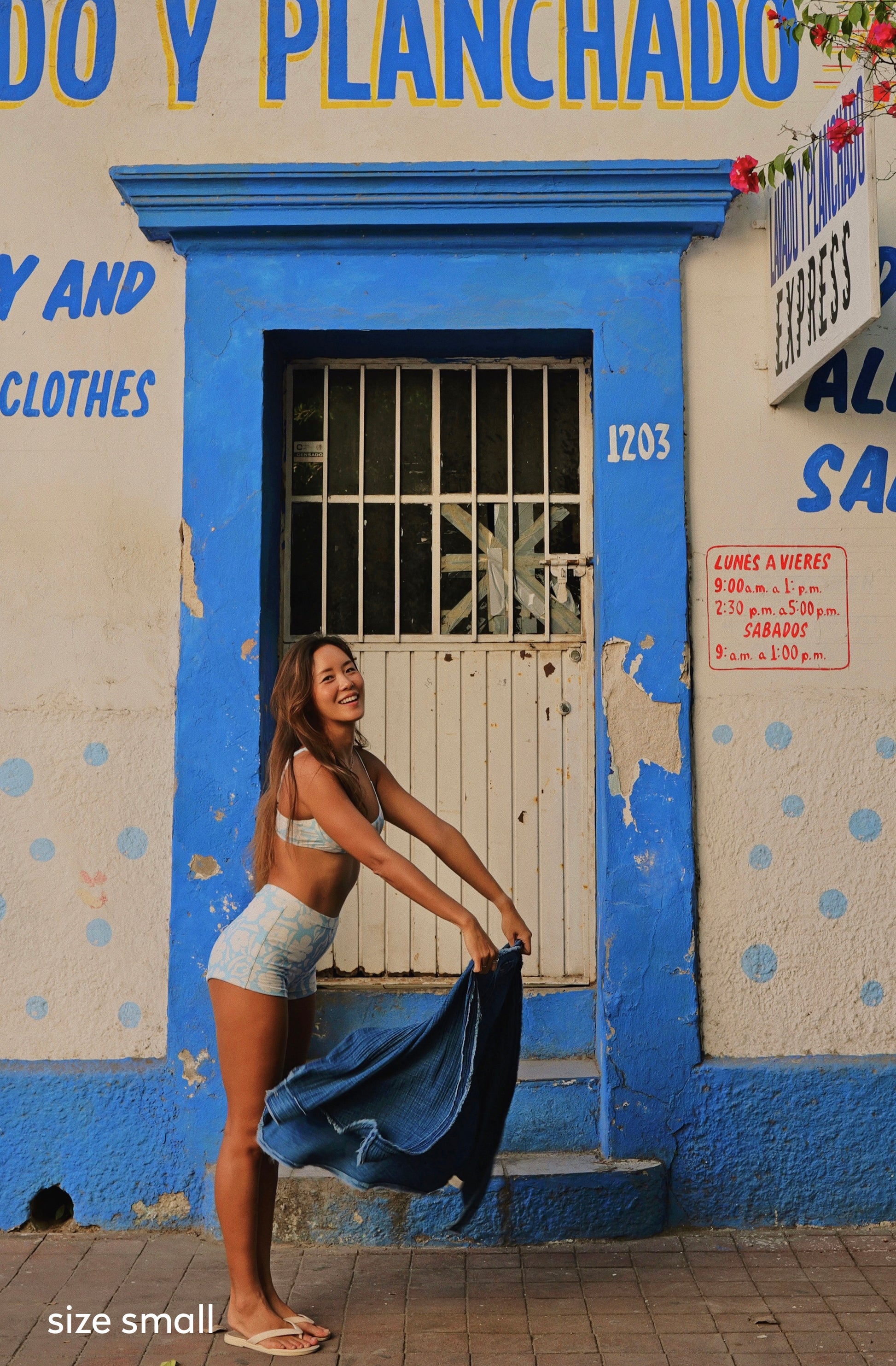 A woman stands in front of a blue and white storefront wall, holding up a blue denim jacket. She is wearing a light beige bikini top and matching light denim shorts.