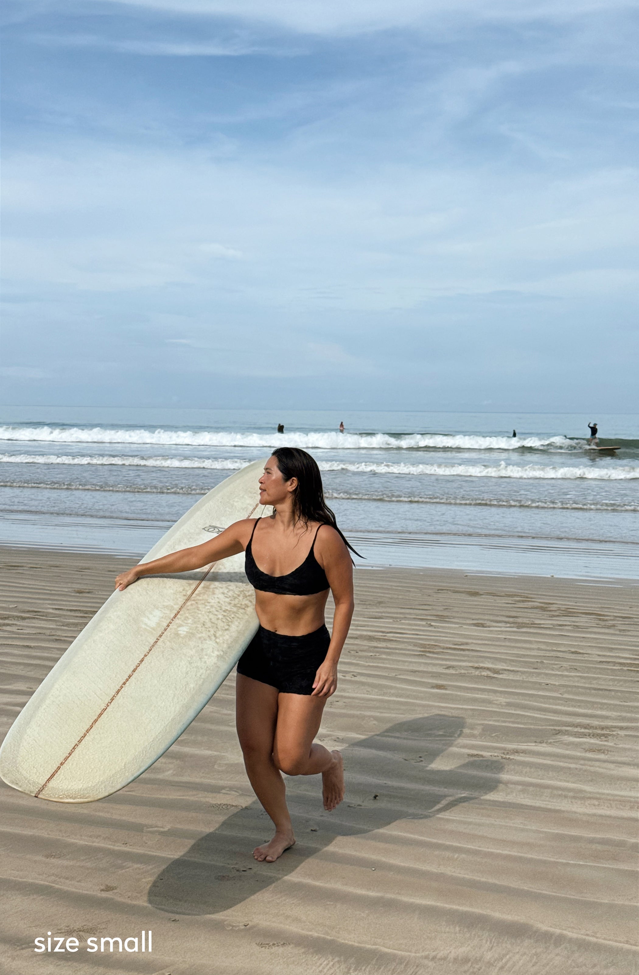 A woman carrying a white surfboard walks on a sandy beach wearing a black two-piece swimsuit, with ocean waves and a blue sky in the background.