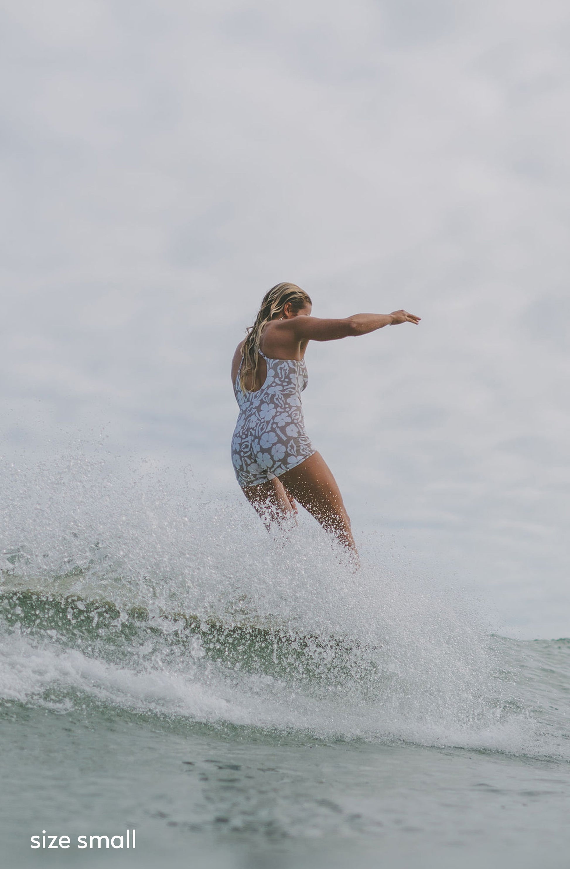 A woman surfing on a small ocean wave, wearing a light-colored patterned one-piece swimsuit. The background is a cloudy, overcast sky and grayish water, with white sea spray around her.