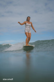 A woman stands on a surfboard on a wave in the ocean, wearing a white one-piece swimsuit with a gray abstract pattern. The sky is cloudy, and the water is a muted blue-green.