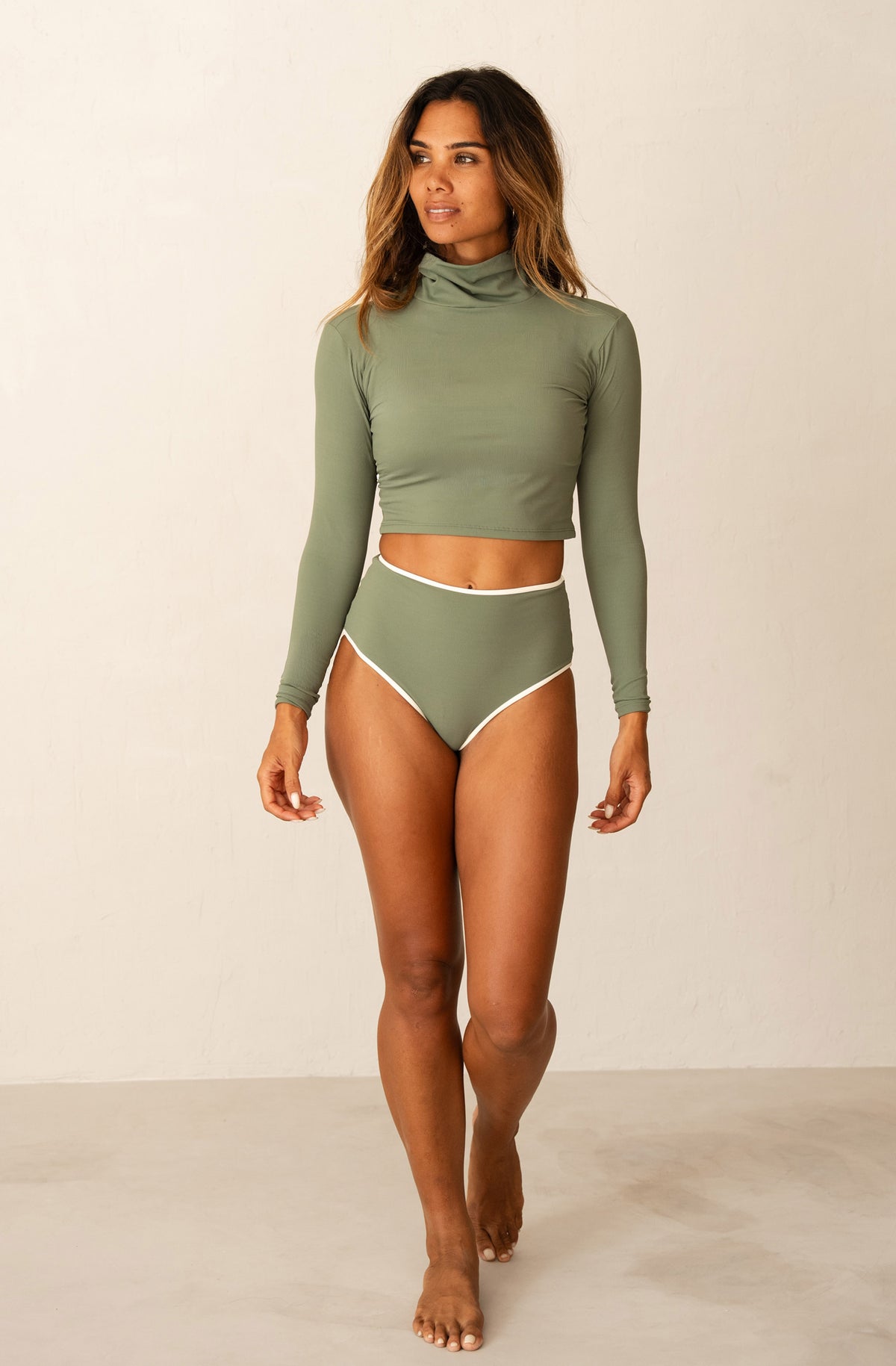 A woman modeling a two-piece swimwear set consisting of an olive green long-sleeve mock neck crop top and matching high-waisted bikini bottoms with white trim, set against a plain off-white studio background.