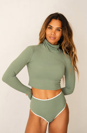 A woman modeling a sage green two-piece swimsuit or lounge set featuring a long-sleeve mock neck crop top and matching high-waisted briefs with a thin white trim, set against a plain off-white background.