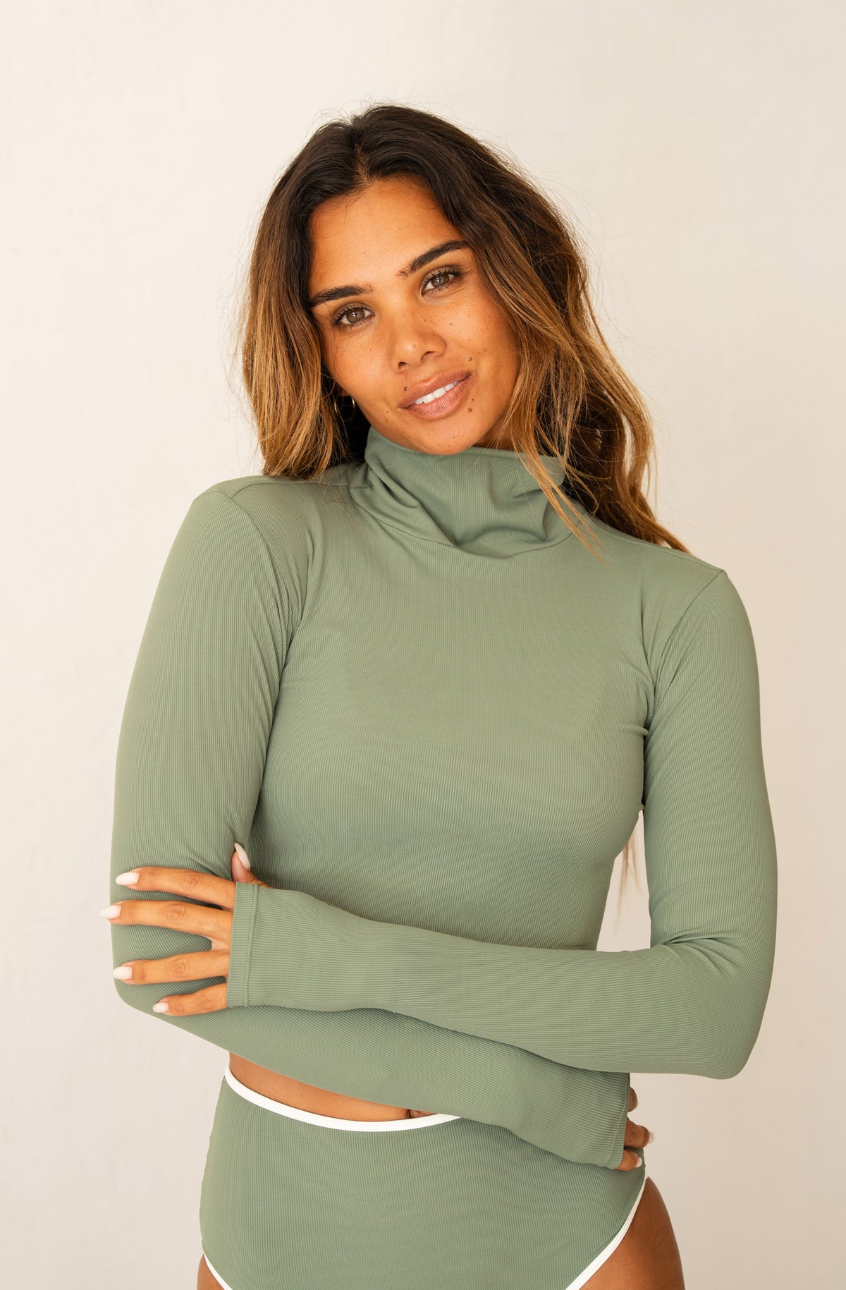 A woman modeling a sage green long-sleeve mock neck crop top and matching high-waisted shorts against a plain off-white background.