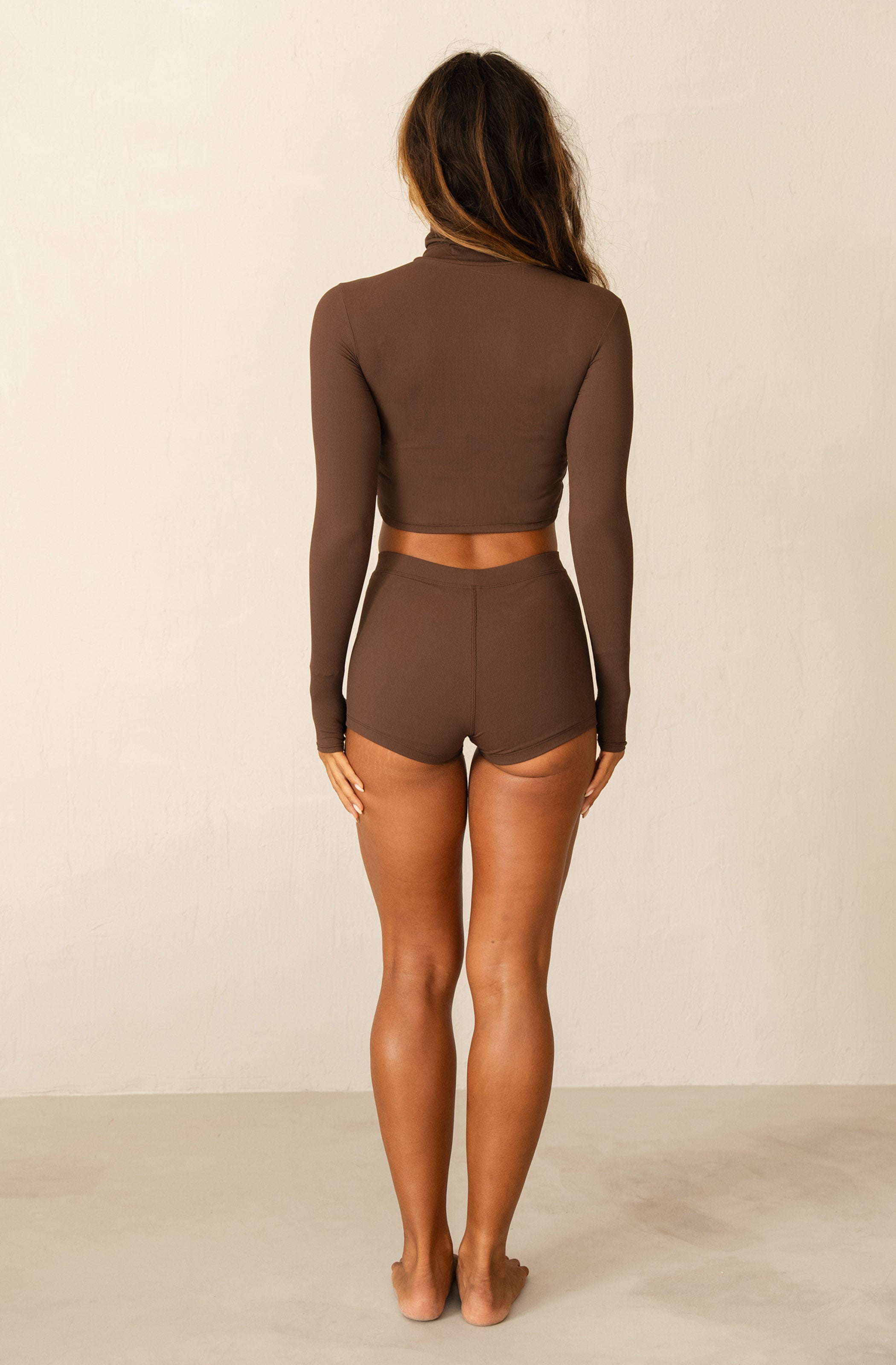 A woman modeling a matching two-piece brown activewear set consisting of a long-sleeve mock neck crop top and high-waisted shorts, set against a plain off-white studio background.