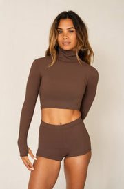 A woman modeling a matching brown ribbed two-piece set consisting of a long-sleeve cropped mock neck top and high-waisted shorts, against a plain light beige studio background.