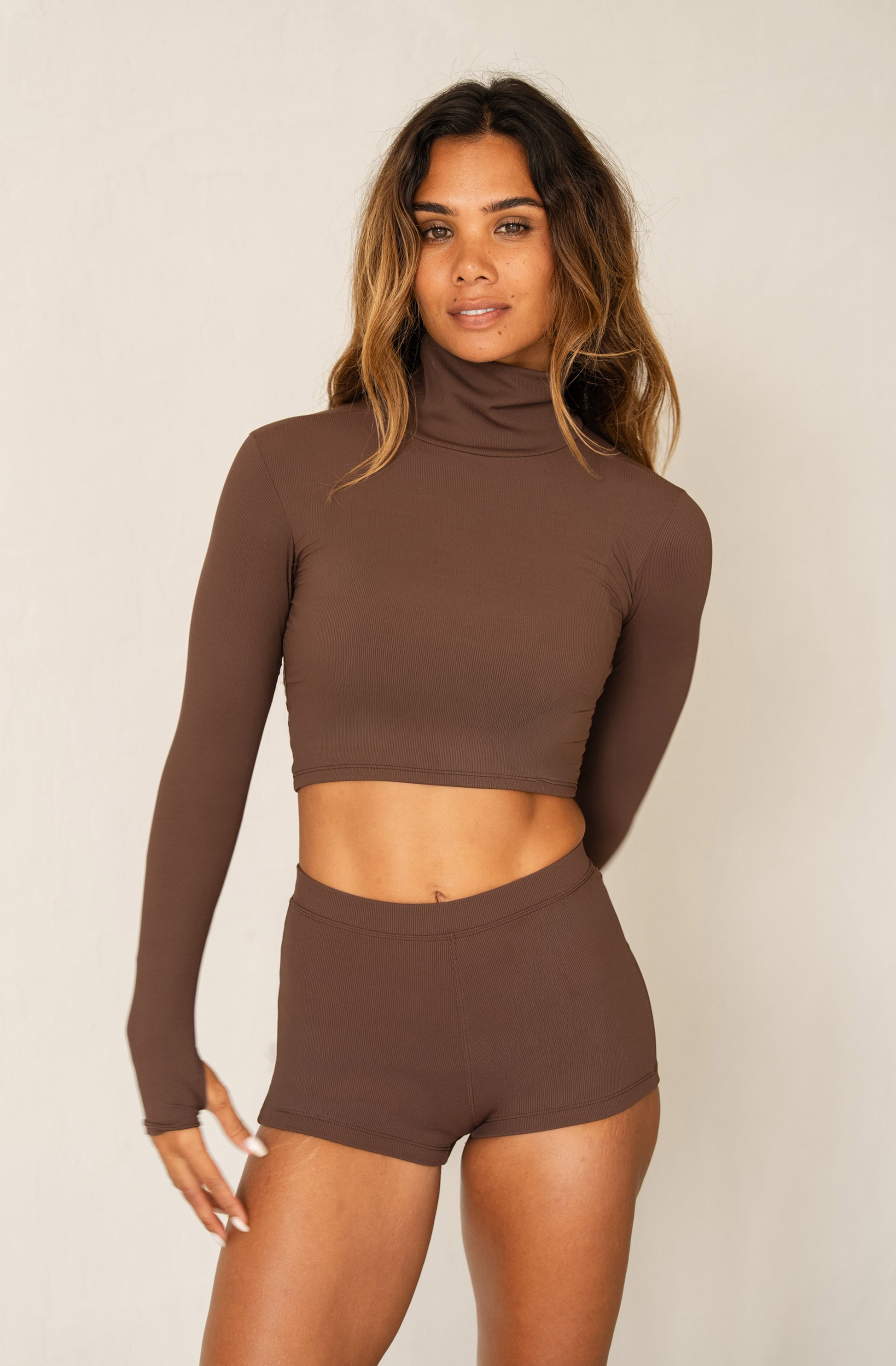 A woman modeling a matching brown ribbed two-piece set consisting of a long-sleeve cropped mock neck top and high-waisted shorts, against a plain light beige studio background.