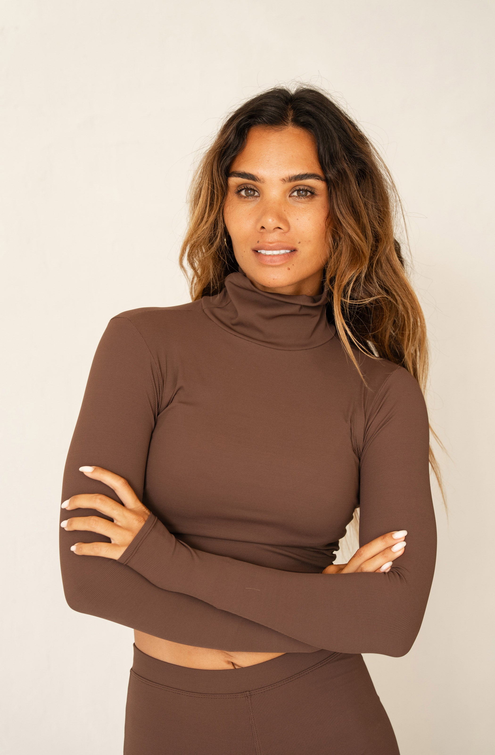 A woman modeling a matching two-piece brown activewear set consisting of a long-sleeve mock neck crop top and high-waisted shorts, set against a plain off-white studio background.