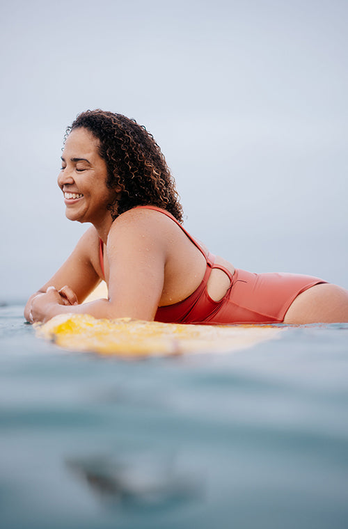 BIPOC woman smiling wearing the Paris One Piece surf swimsuit as she's laying on her surfboard in the ocean. 
