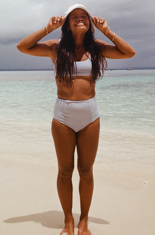 BIPOC woman standing on the beach wearing the Jane surf bikini top and Alice Surf bikini bottom wearing a sun hat smiling looking up at the sky 