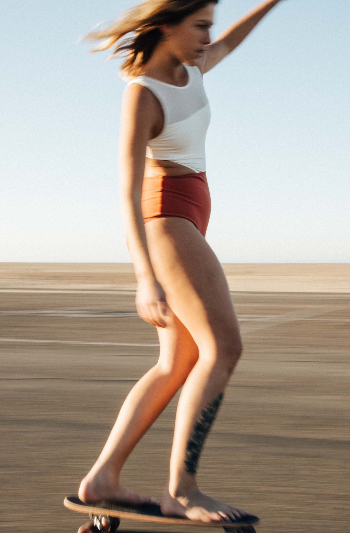 Shot of a woman skating wearing a one piece surf swimsuit in ivory/burnt sienna.