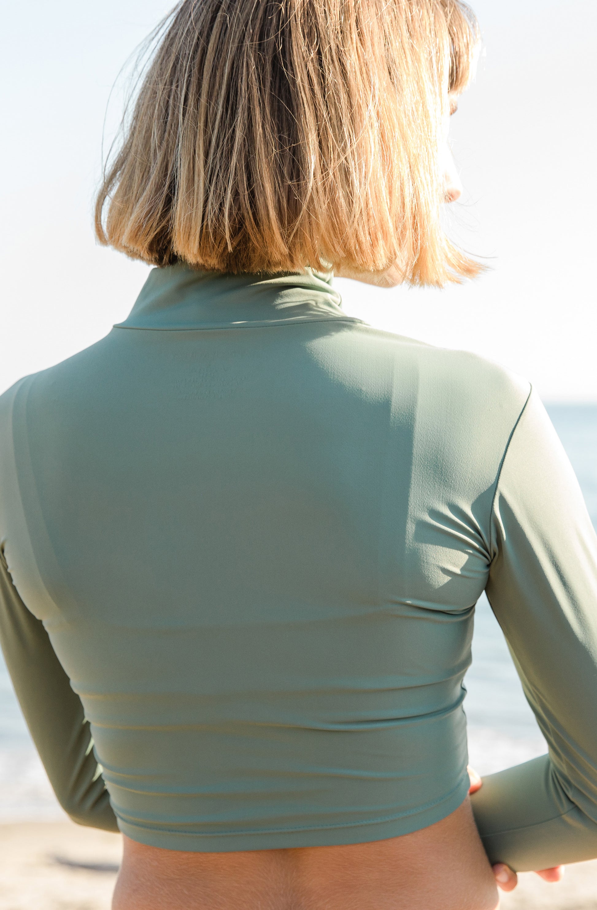 Close back shot of a woman at the beach modelling a cropped rash guard and a surf bikini bottom in army green.