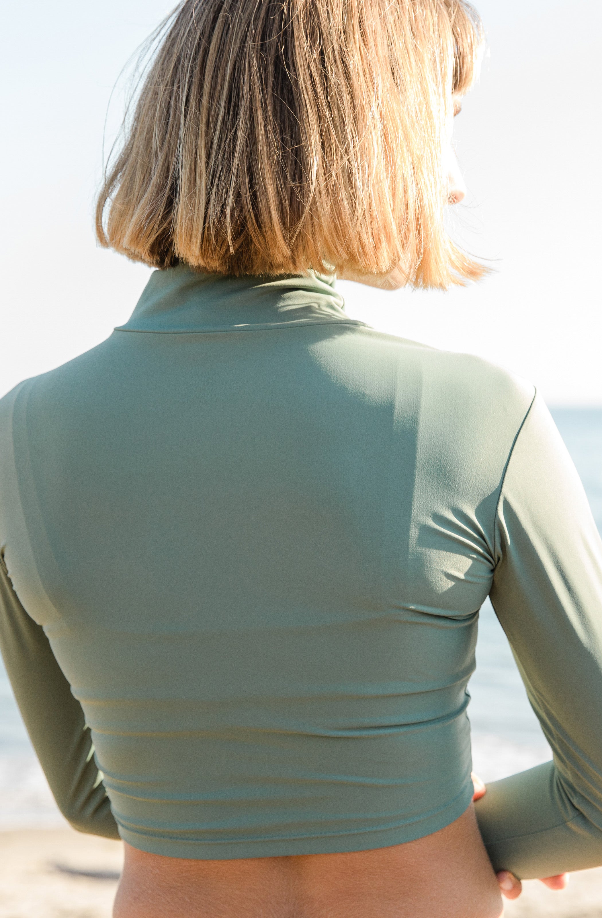 Close back shot of a woman at the beach modelling a cropped rash guard and a surf bikini bottom in army green.