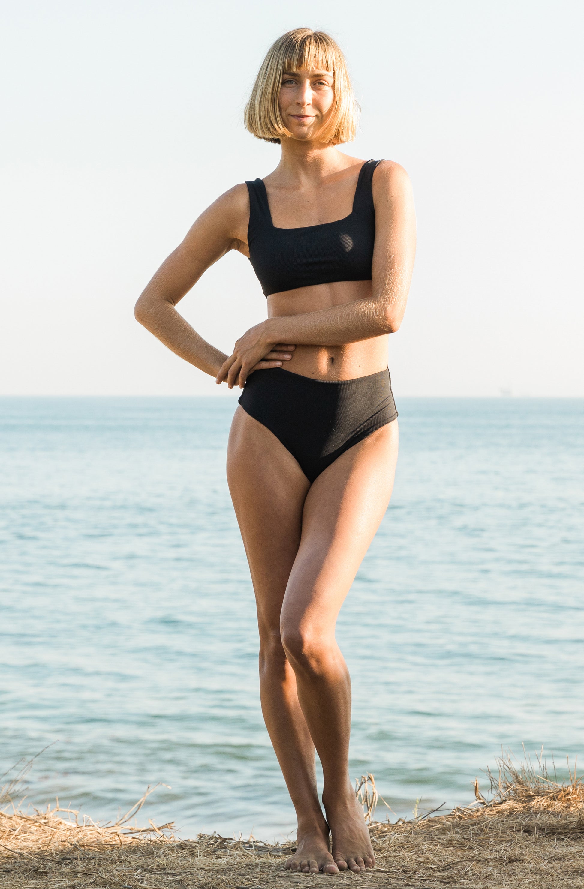 Front view of a woman at the beach modelling a ribbed surf bikini top and a ribbed high-waisted bikini bottom in black.