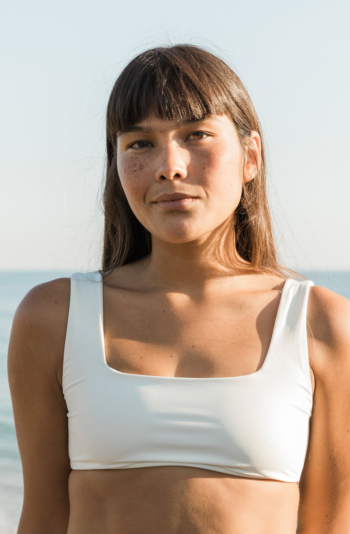 Front view close shot of a woman in front of the ocean wearing a surf bikini top in ivory.