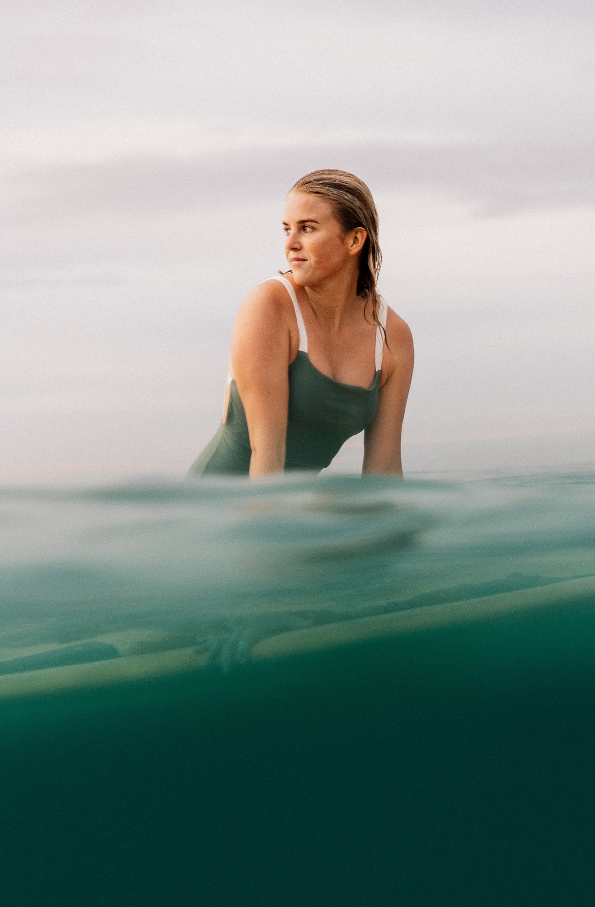 Shot of a woman on a longboard wearing a one piece surf swimsuit in army green.