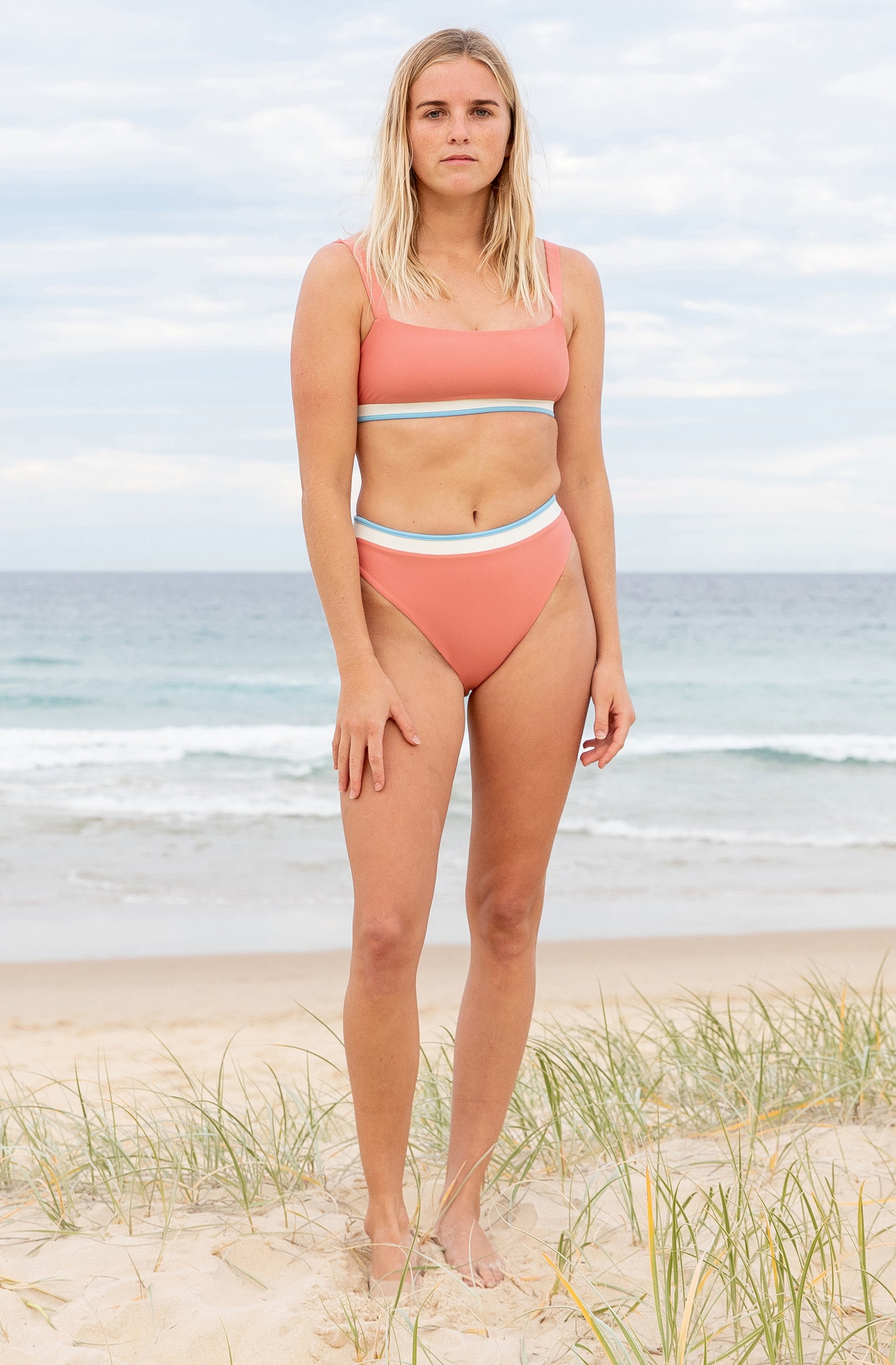 Front view of a woman at the beach modelling a surf bikini top and a surf bikini bottom in vintage blush.